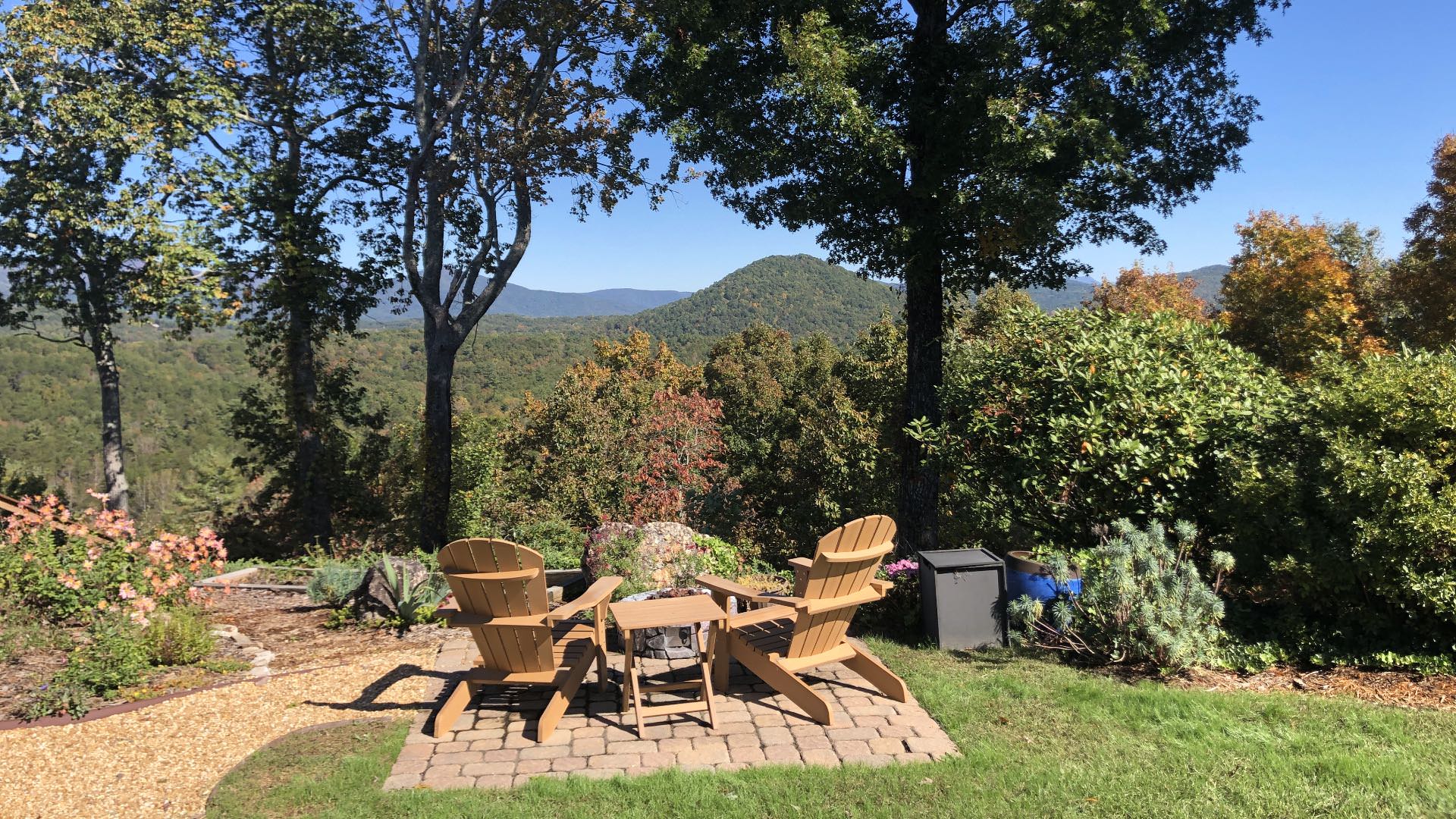 Two light tan Adirondack chairs on brick patio with fire pit surrounded by flower garden, bushes, and trees looking out to tree-covered hills