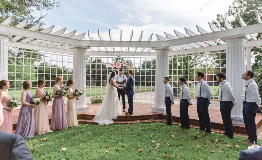 Wedding party standing on grass near large white pergola with big white columns