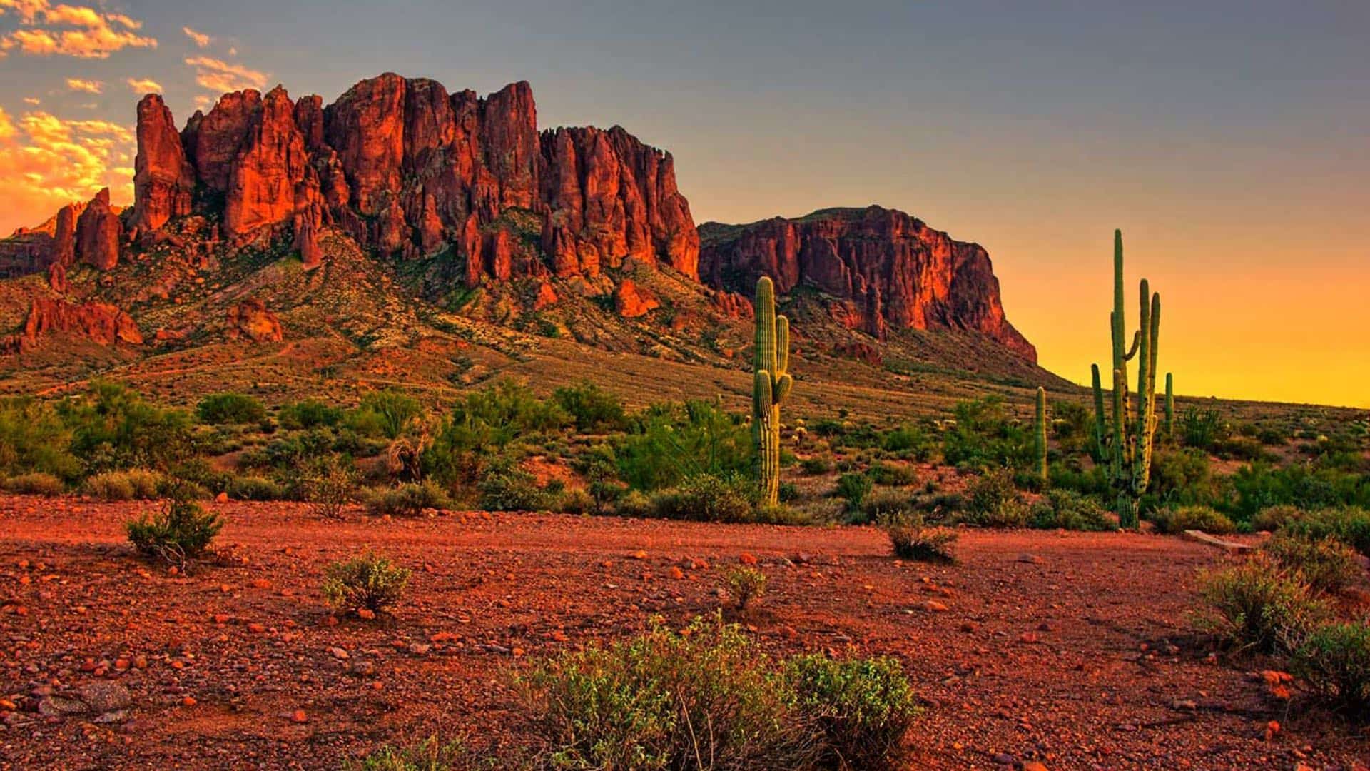 Tall and jagged red mesas surrounded by red dirt, green shrubs, and tall cacti