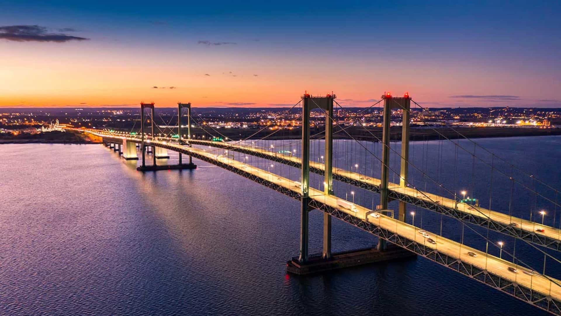 Large lighted up bridge across a large body of water with a city in the background at dusk