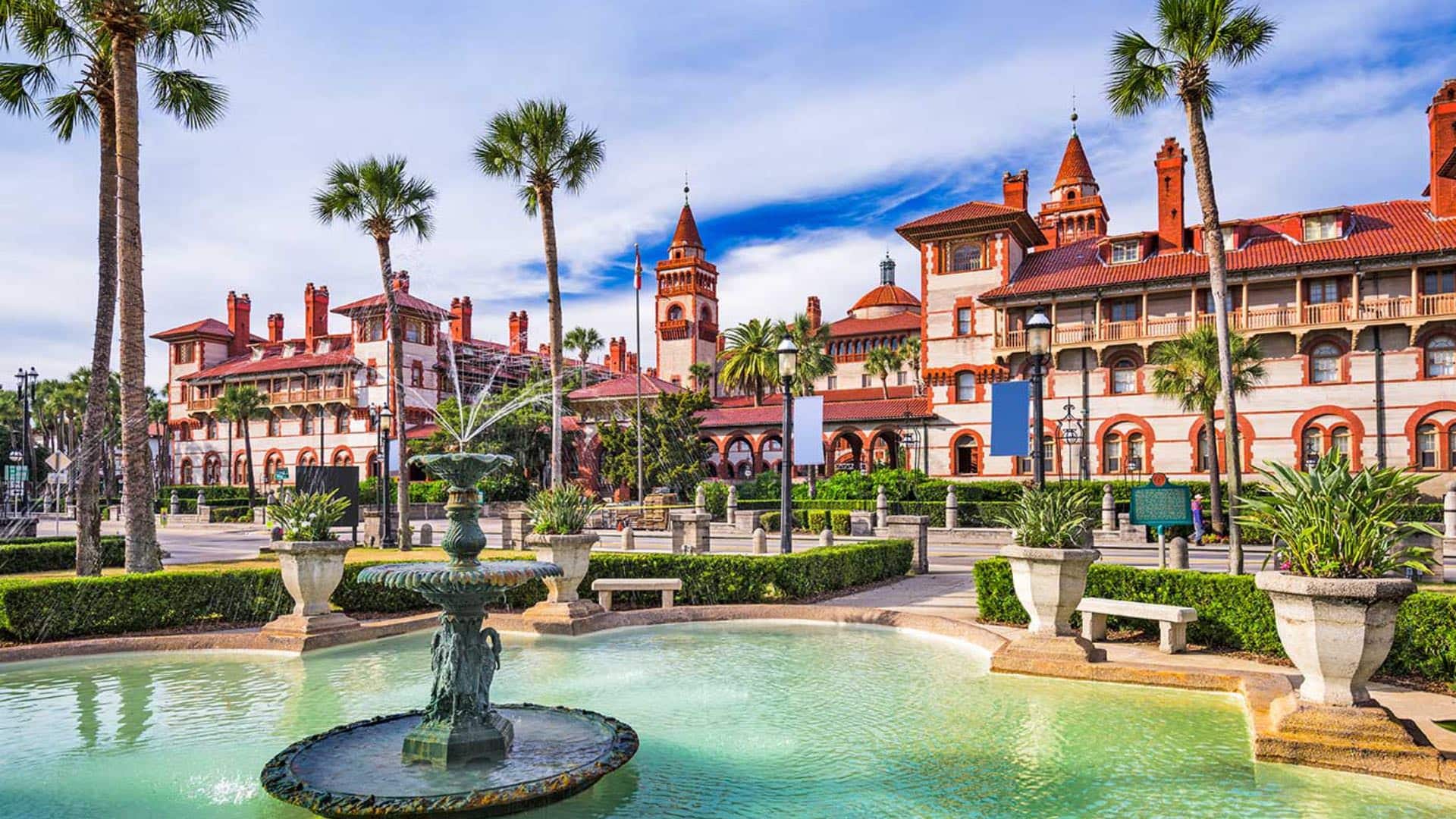 Large property painted light tan with bright orange trim, palm trees, green shrubs and a water fountain across the street