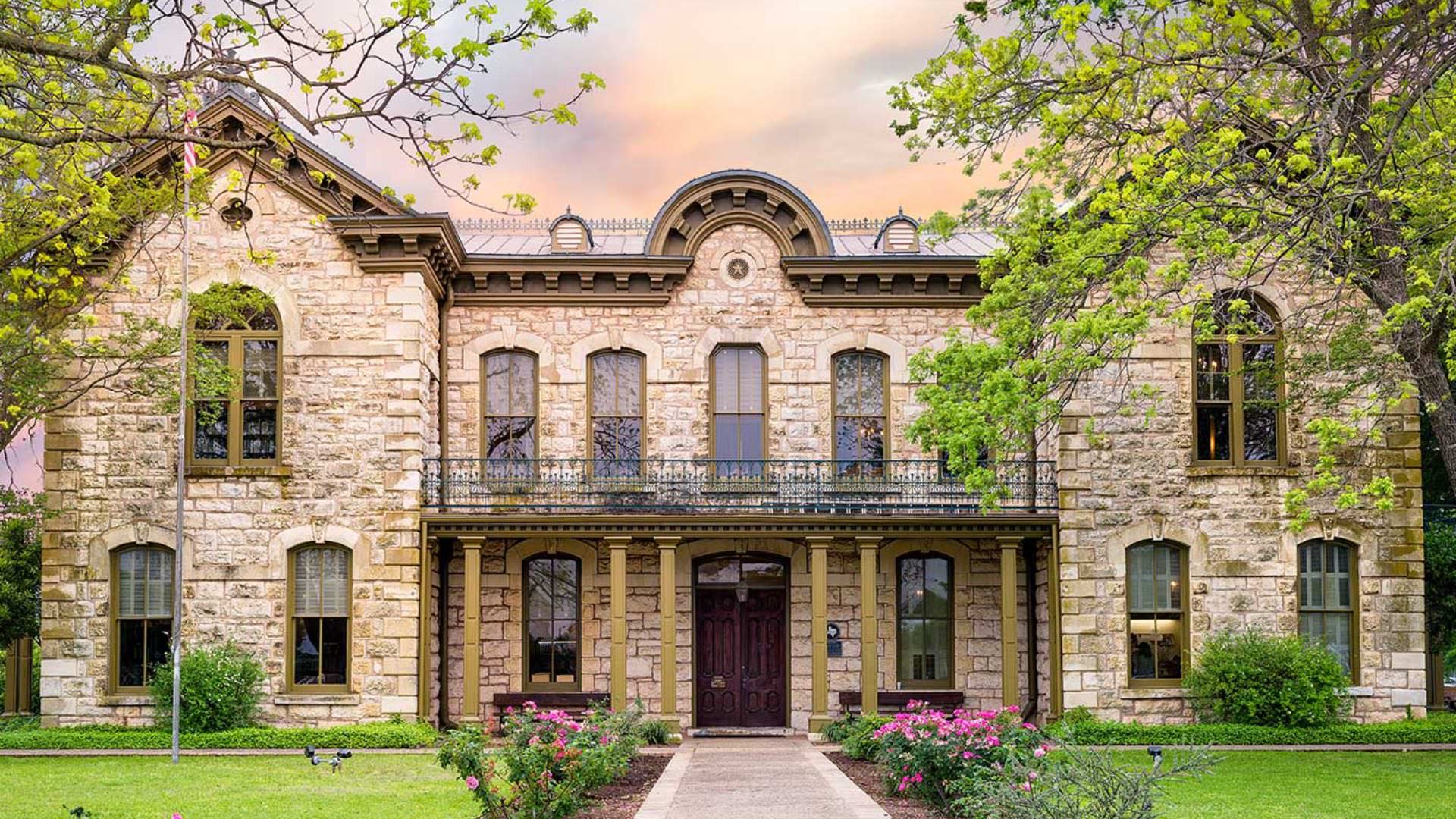 Exterior view of a property with stone brick walls, olive green trim, green grass, and sidewalk flanked by green bushes with pink flowers