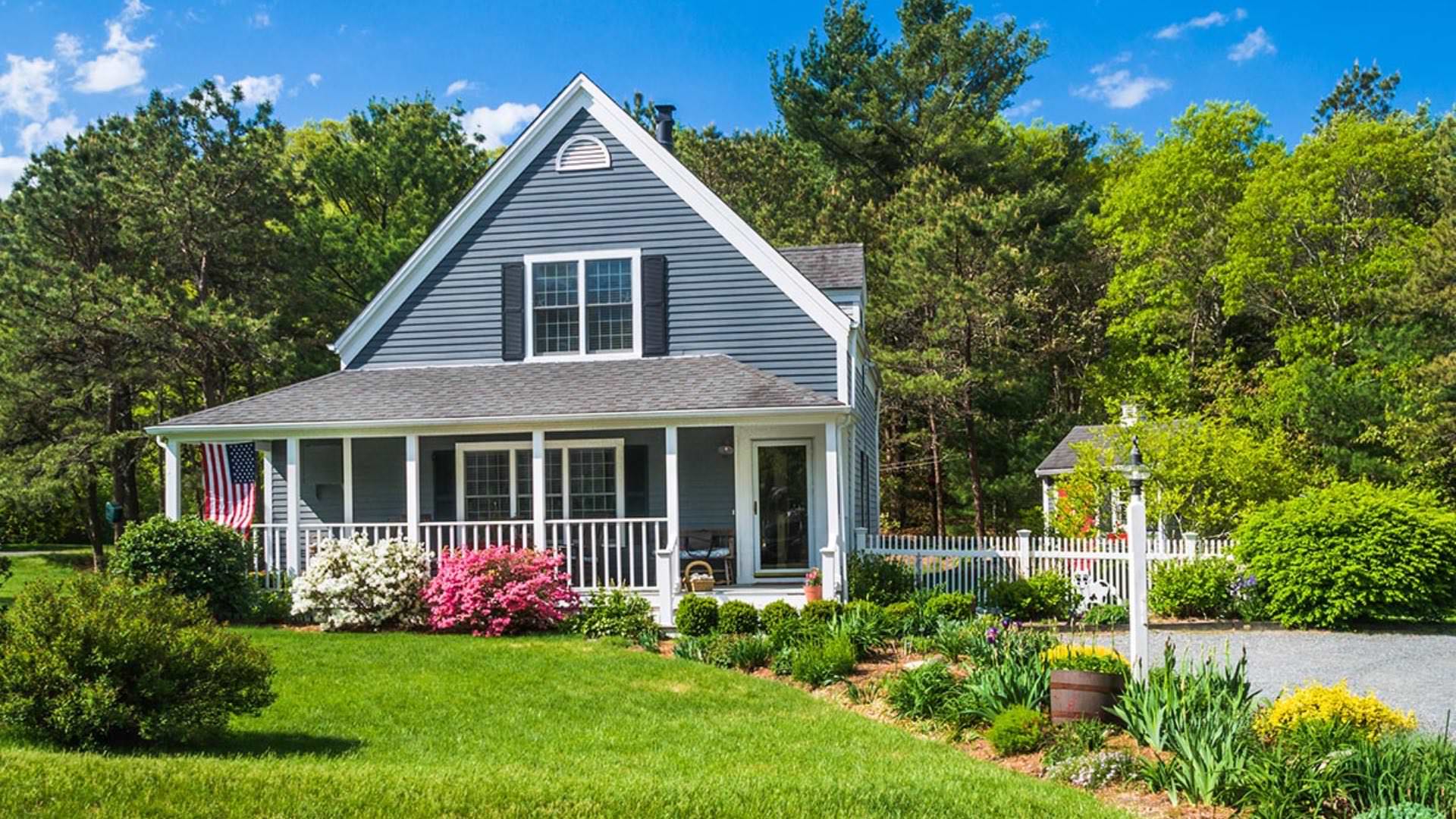 Exterior view of property painted gray blue with white trim, navy blue shutters, front porch, and surrounded by green trees