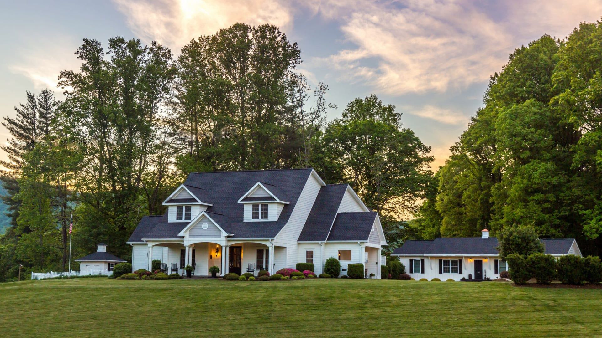 Exterior view of property painted white with white trim and surrounded by green bushes, large green lawn, and tall green trees