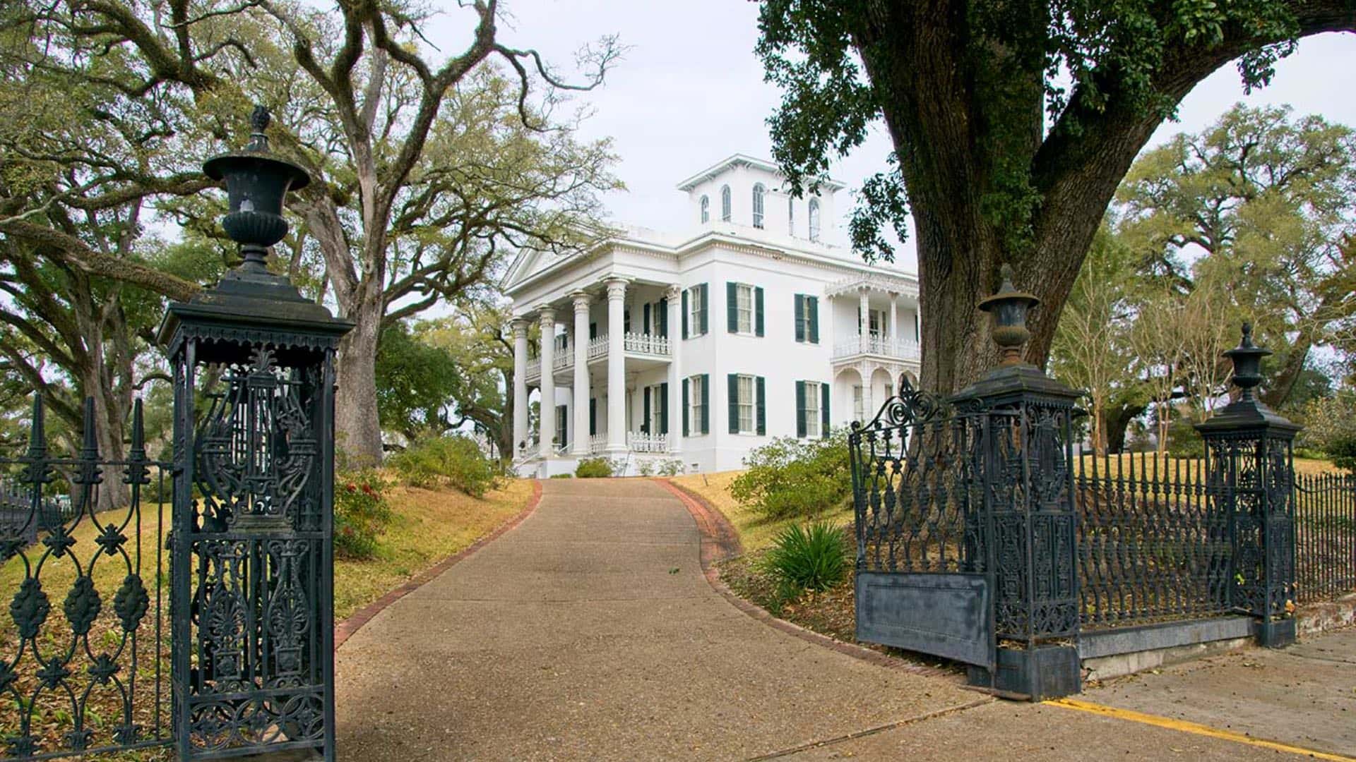 Exterior view of a property painted white with green shutters, large white columns, large trees and surrounded by an antique wrought iron fence