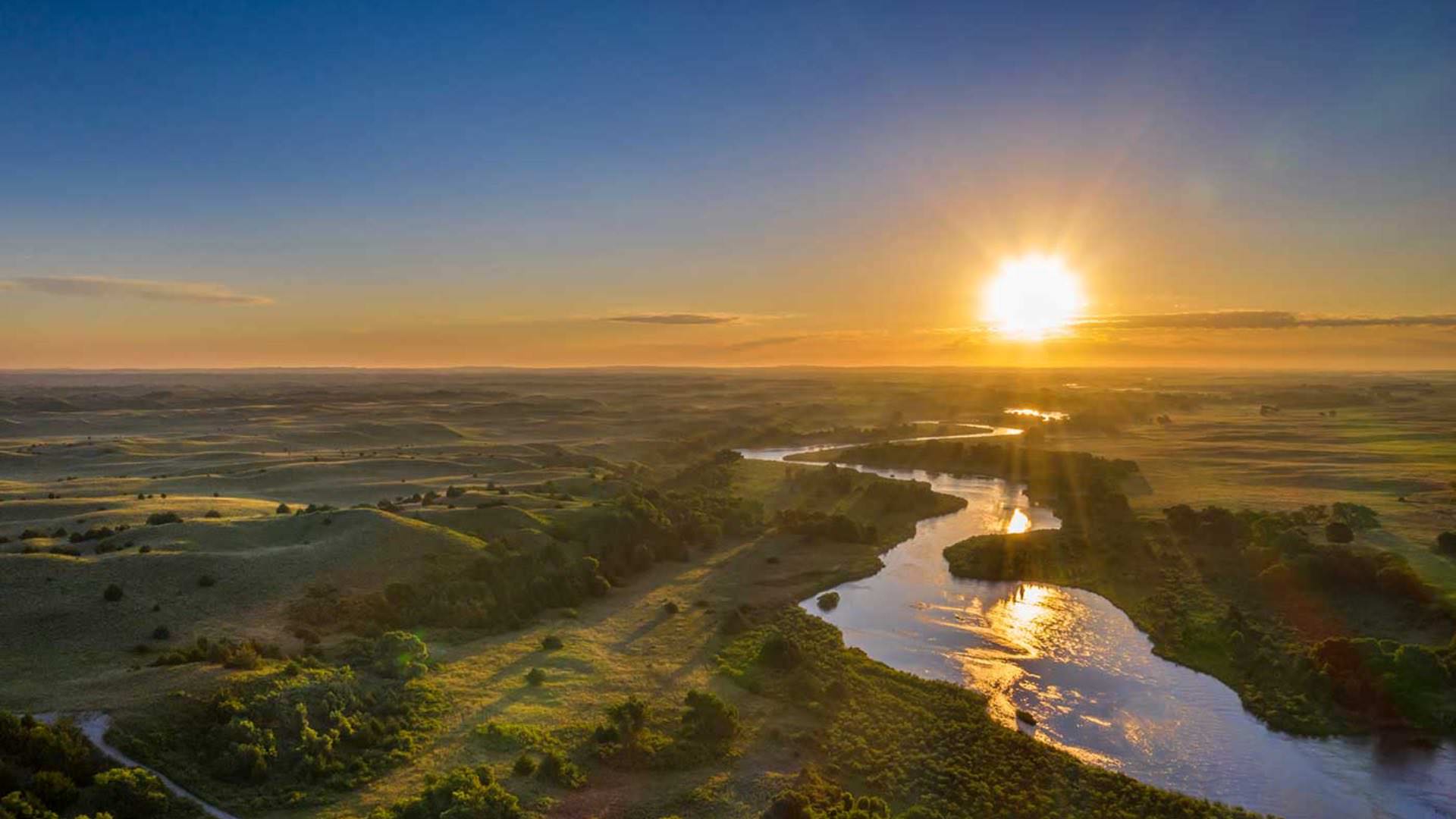 Aerial view of a winding river surrounded by green grass, rolling hills, and trees with the setting sun in the background