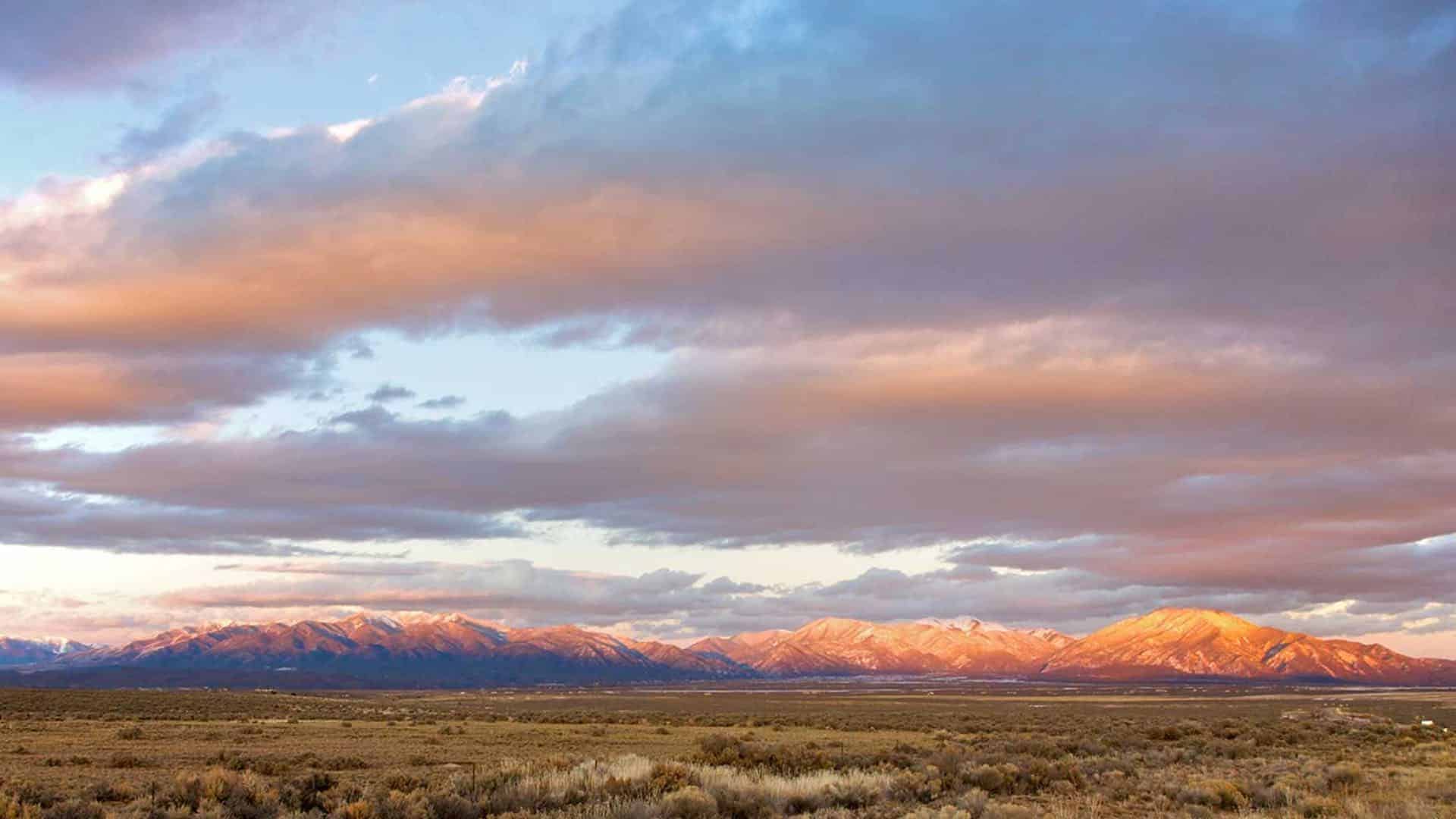 Plains shaded in browns and greens with mountain range and sky full of clouds in the background