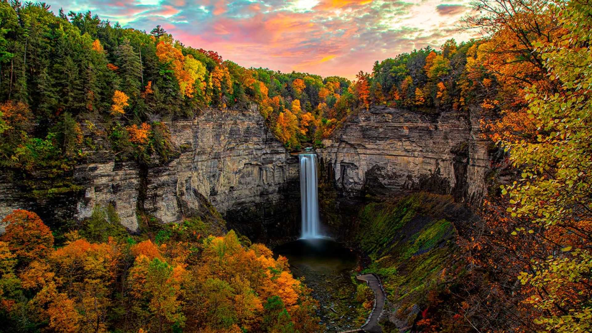 Small waterfall coming out the side of a curved cliff surrounded by green, orange, and yellow trees