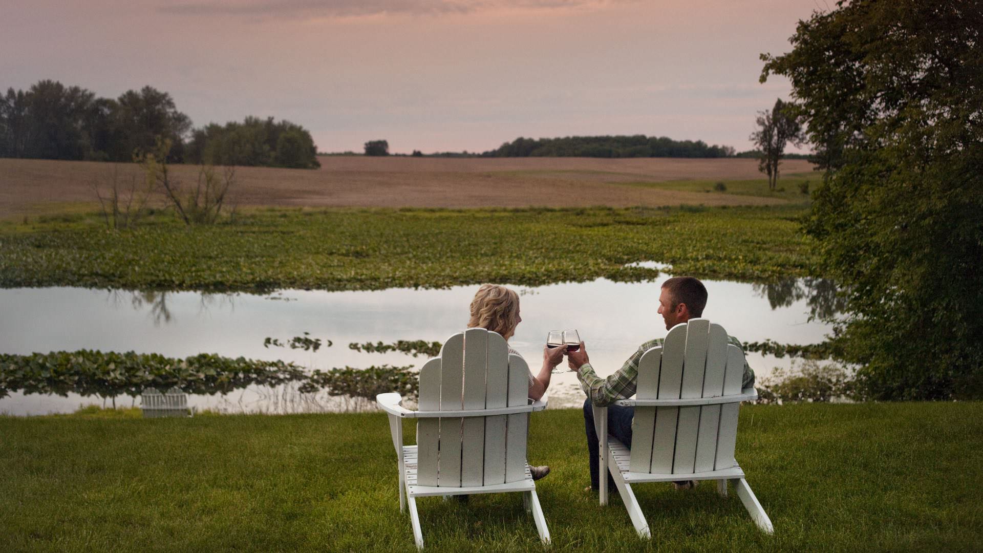 Two people sitting on white Adirondack chairs holding wine glasses looking at each other with a pond in the background