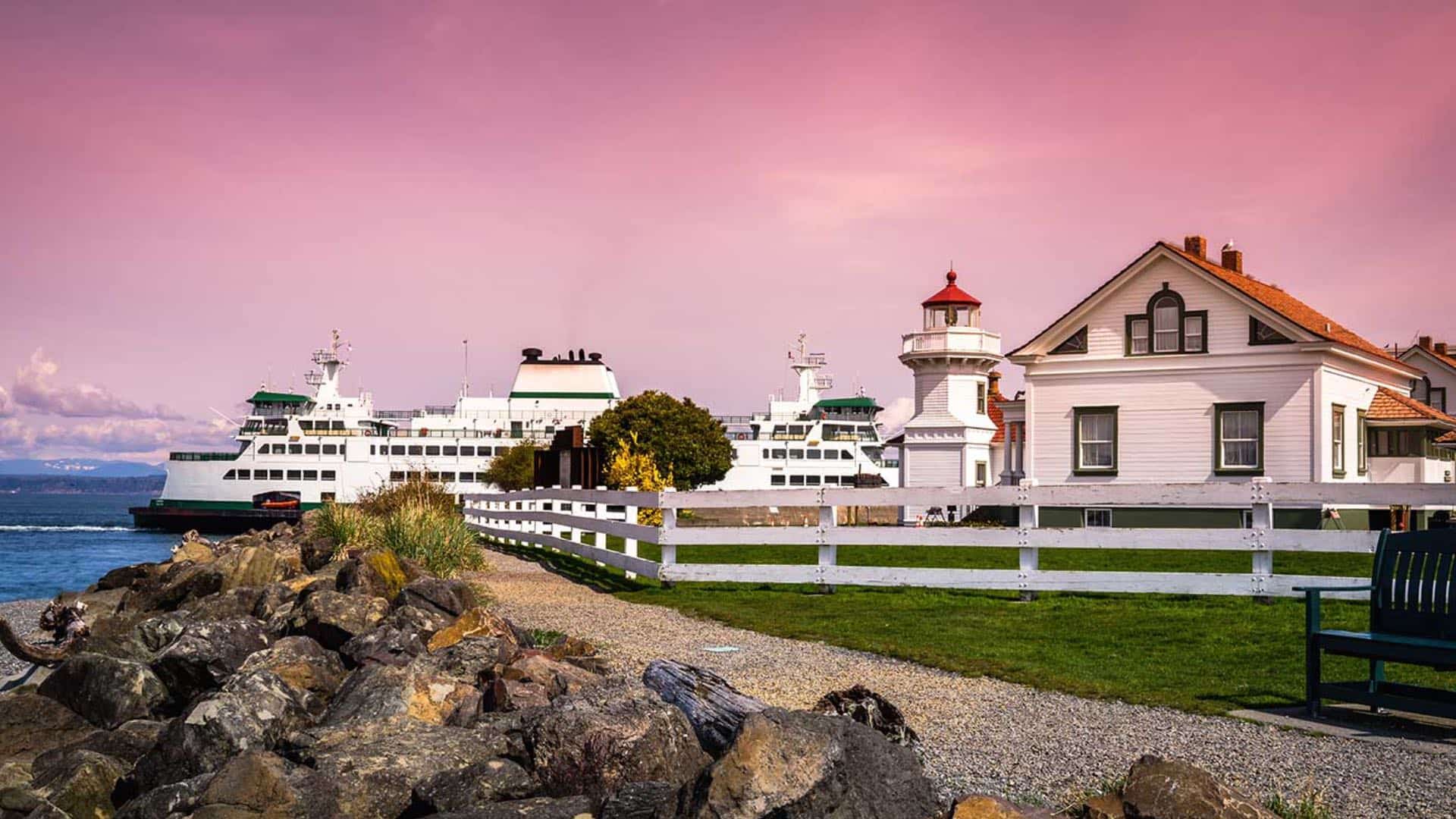 White lighthouse and house surrounded by green grass and white fence with large white boat in the background