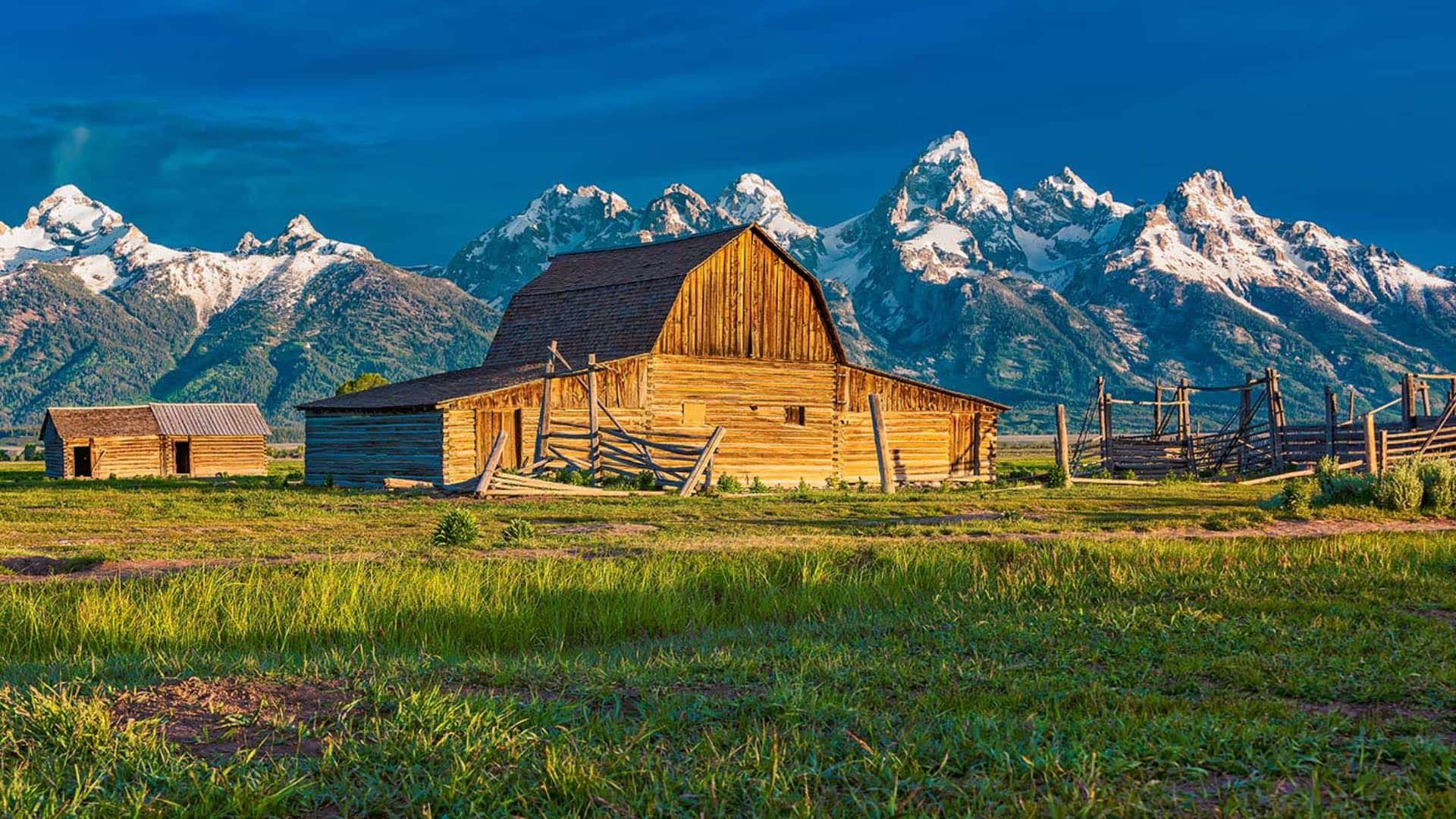 Old wooden barn surrounded by green grass with jagged snow-capped mountains in the background