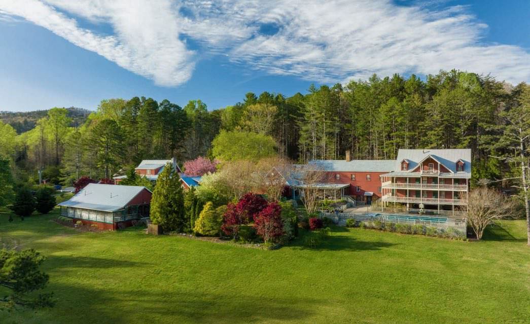 Exterior view of three-level property with red brick, white trim, white wrap around porch and balconies, pool, and surrounded by large green lawn and green trees