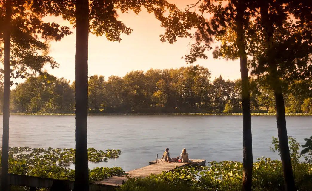 Two people sitting on a dock looking out over a lake surrounded by large trees